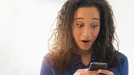 Woman early twenties, looking thrilled with/surprised by what she sees on her phone. 