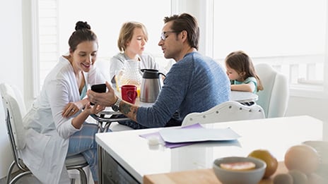 Couple sitting at kitchen table with their 2 young children. Father is showing mother something on his phone. 